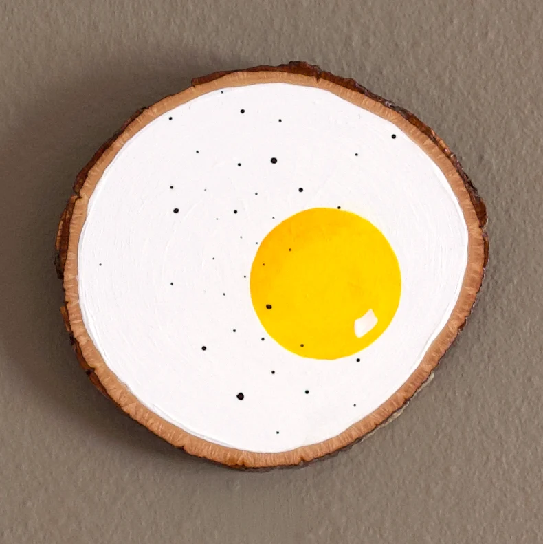 A slice of wood is painted to resemble a fried egg with a yellow yolk and white background.