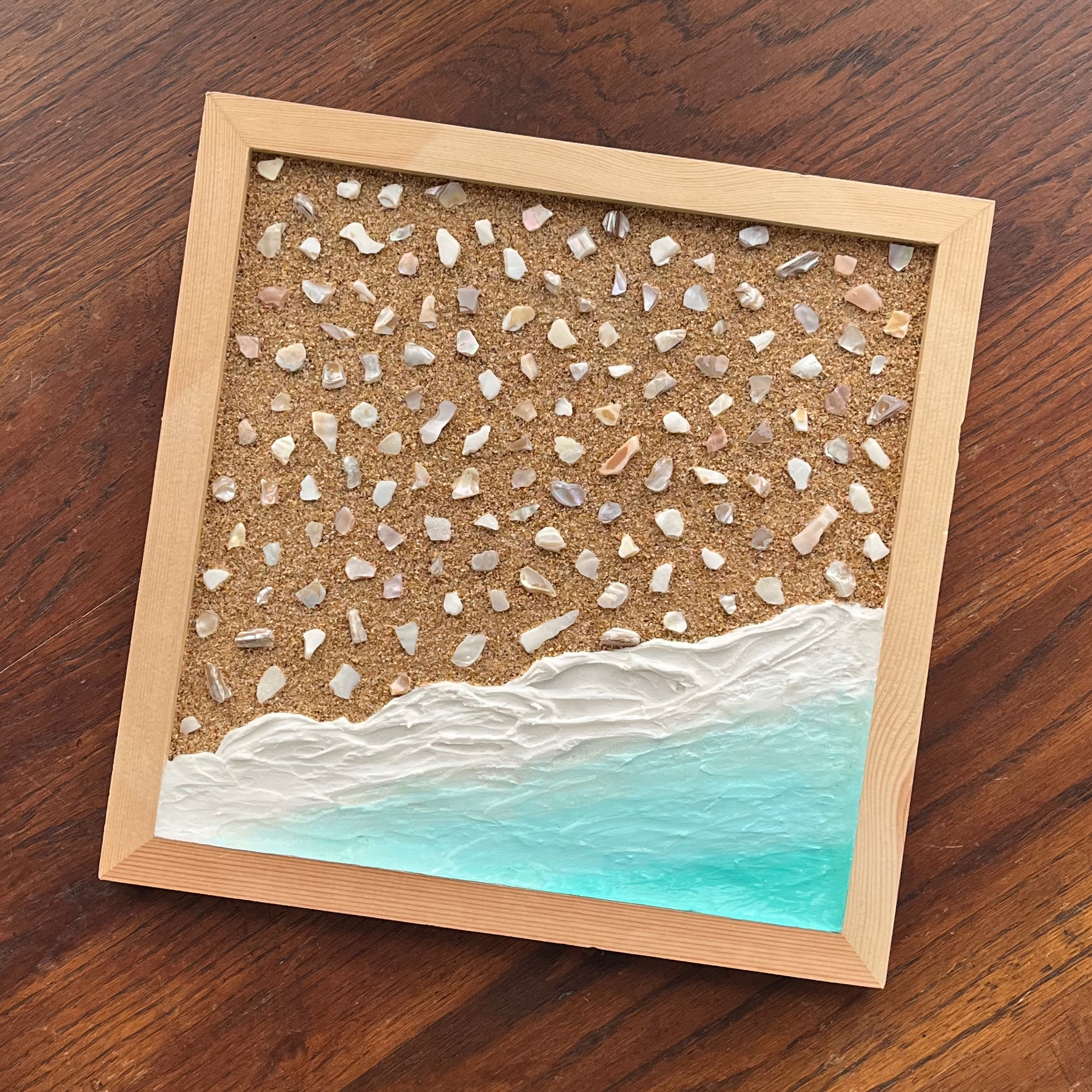 A framed artwork creatively depicts a beach scene with textured sand and vibrant blue waves.