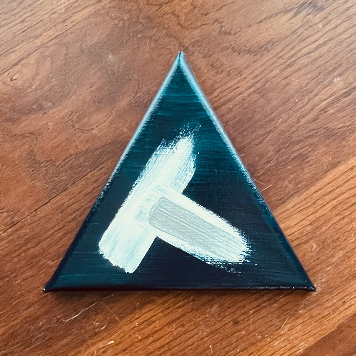 A small triangular canvas features an abstract painting with dark teal and white brushstrokes, set against a wooden surface.