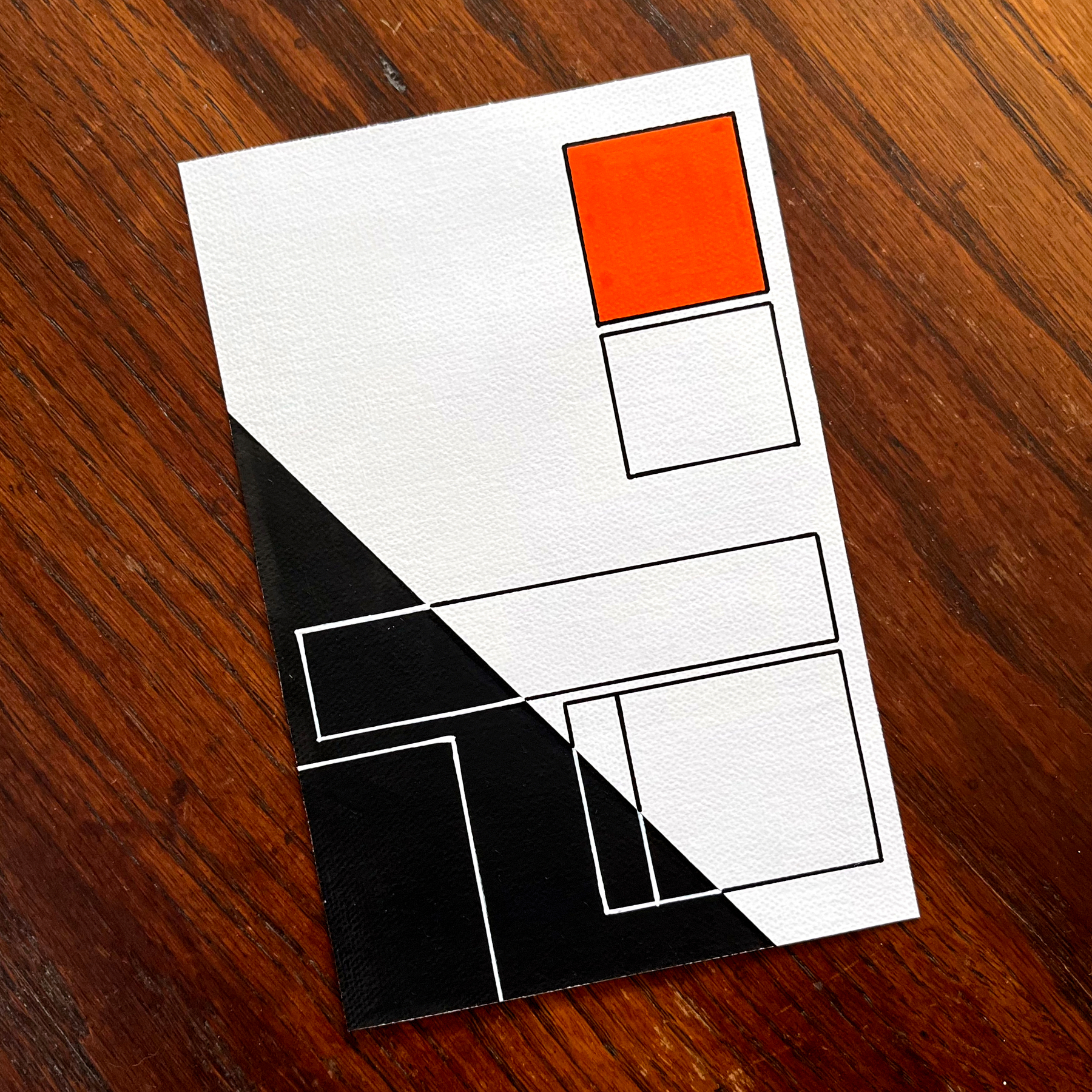 A minimalist abstract artwork features a geometric arrangement of rectangles in black, white, and orange on a wooden surface.